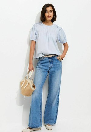 Kobieta ubrana w jasnoniebieską bluzkę z bufkami i szerokie dżinsy, trzymająca beżową plecioną torebkę, stojąca na białym tle.