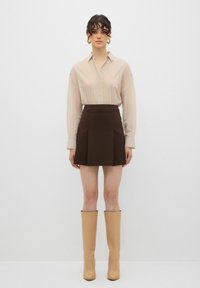 Blusa plissettata beige con bottoni in evidenza, abbinata a una mini gonna marrone. Il modello indossa stivali beige fino al ginocchio con una finitura liscia.