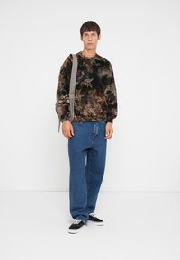 Bruine en groene camouflage-patroon trui met elastische manchetten en een relaxte pasvorm, gecombineerd met blauwe rechte spijkerbroeken en zwarte sneakers.