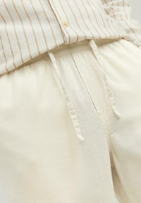 Light beige linen joggers feature an elastic waistband with a drawstring and side pockets. Smooth texture with a relaxed fit.