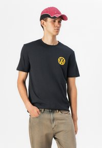 Sort bomulds t-shirt med et gult Volkswagen-logo på brystet, parret med lysebrune denimjeans og en lyserød baseballkasket.