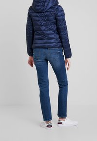 Person iført en marineblå pufferjakke, blå jeans og hvide sneakers med sølv- og røde detaljer, stående mod en ensfarvet grå baggrund.