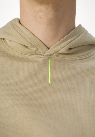 Beige kapucnis pulóver struktúrált felülettel és kontrasztos neon zöld függőleges varrással a nyakkivágásnál.