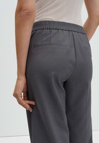 Grijze, op maat gemaakte broek met een elastische tailleband, twee achterzakken en een gladde textuur, getoond vanuit een zijaanzicht.