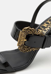 Sandalo con tacco alto in vernice nera, caratterizzato da una fibbia dorata decorativa con accenti floreali e un design a fascia. Si allinea con lo stile del calzature formali.