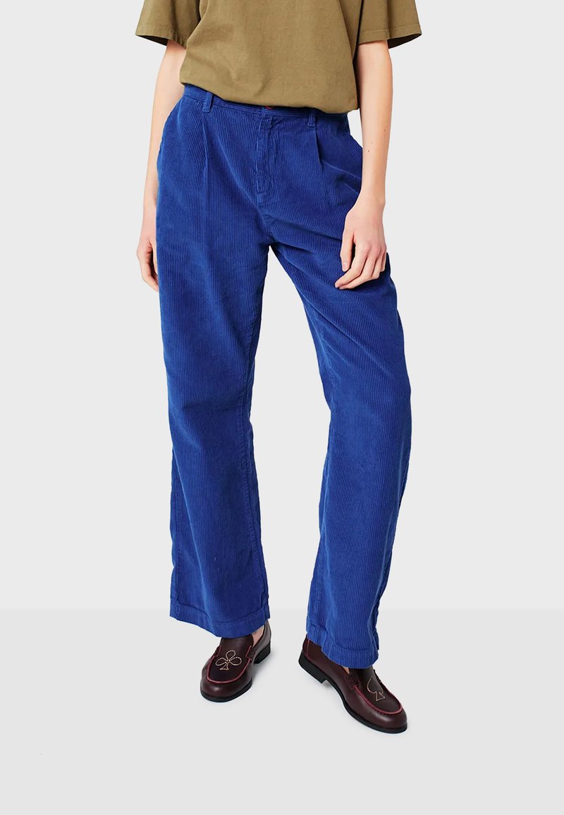 Persona che indossa pantaloni in corduroy blu a vita alta, una maglietta verde oliva infilata dentro e scarpe in pelle marrone scuro con ricamo a tema carte da gioco.