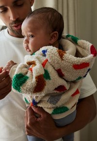 Veste à capuche en polaire crème avec des motifs géométriques colorés. Elle présente un tissu doux et texturé, des manches avec poignets et une capuche spacieuse.
