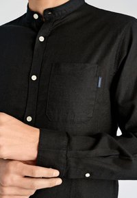 Czarna koszula na guziki z kołnierzykiem stojącym, z kieszenią na piersi, białymi guzikami i fakturowaną tkaniną.