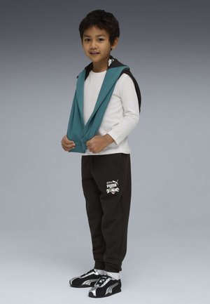 Ragazzo che indossa una maglietta bianca a maniche lunghe, pantaloni della tuta neri Puma, scarpe da ginnastica nere e argento, e tiene una felpa con cappuccio verde acqua sulle spalle.