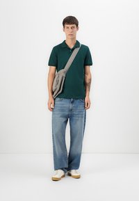 Sötétzöld pólóing, laza kék farmernadrág, szürke crossbody táska, fehér sportcipők bézs talppal. Egyszerű, letisztult dizájn, laza fazon.