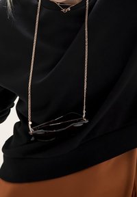 Sweat-shirt noir à coupe ample, avec une chaîne dorée tenant des lunettes de soleil, associé à une jupe marron. Texture douce et design minimaliste.