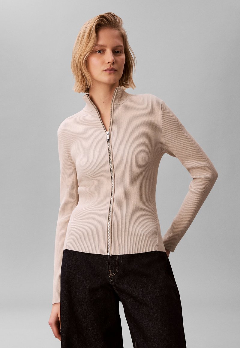 Suéter beige claro acanalado con cuello alto y cremallera, corte entallado y mangas largas, combinado con pantalones vaqueros oscuros de tela texturizada.