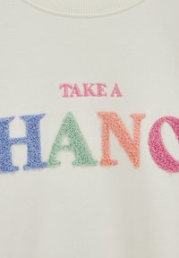 Balts audums ar izšūtu tekstu "UZŅEM PELNIS" rozā, zilās, zaļās, oranžās un magentas strukturētās burtiņās.