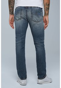 Jeans azul oscuro con un acabado desgastado, que cuentan con dos bolsillos traseros y detalles de costura visibles. Llevados con zapatillas deportivas blancas.