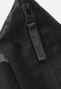 Svart vanntett bag laget av glatt materiale med glidelås. Har et strukturert stripe og et merke som viser "RAINS."
