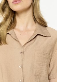 Light beige short-sleeve button-up shirt with a collar, featuring a pocket on the left side and smooth, textured fabric.