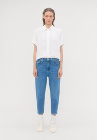 Žena stojaca čelom s bielou košelou s krátkym rukávom, modrými džínami, bielymi ponožkami a bielymi teniskami na jednoliatej pozadí.