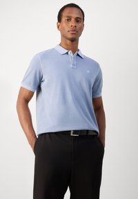 Camisa polo azul claro con cuello y mangas cortas, con un sutil logo en el lado izquierdo del pecho, combinada con pantalones negros y un cinturón.