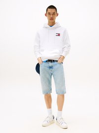 Sudadera blanca con logo, pantalones cortos de mezclilla azul claro, zapatillas blancas con detalles en marino y calcetines blancos. Conjunto casual con un ajuste relajado.