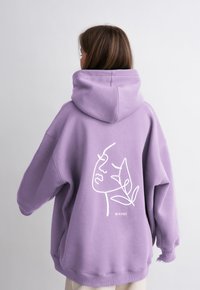 Hoodie oversize in cotone leggero color viola chiaro con una grande grafica bianca outline di un volto e una foglia nella parte posteriore. Presenta un cappuccio spesso e polsini a coste.