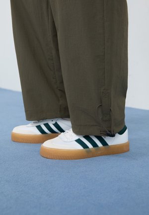 Personne portant des baskets blanches avec des bandes vert foncé et des semelles en gomme, associées à un pantalon ample vert olive sur un sol bleu.