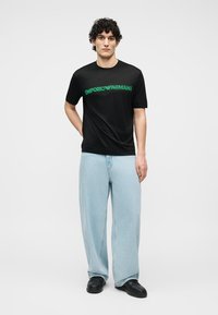 Νέος άνδρας που στέκεται με το ένα χέρι πίσω από την πλάτη, φοράει μαύρο μπλουζάκι Emporio Armani, ανοιχτό μπλε τζιν παντελόνι με φαρδιά πόδια και μαύρα παπούτσια.