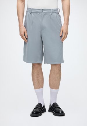 Hombre con pantalones cortos gris claro hasta la rodilla, calcetines blancos y mocasines de cuero negro, de pie contra un fondo blanco liso.