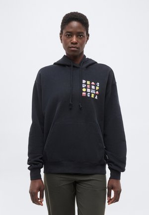 Henkilö pukeutuneena mustaan huppariin, jossa värikkäitä geometrisia tekstejä rinnassa, ja tummanvihreisiin housuihin, seisomassa yksinkertaisen vaalean taustan edessä.