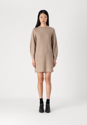 Strikket beige kjole med båthals og lange ermer, med ribbet tekstur og en lett fillete kant. Brukt med svarte sokker og sko.