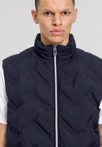 Marineblauer Steppweste mit hohem Kragen, ausgestattet mit einem Frontreißverschluss und einem gesteppten Design. Getragen über einem weißen Hemd. Keine sichtbaren Taschen.