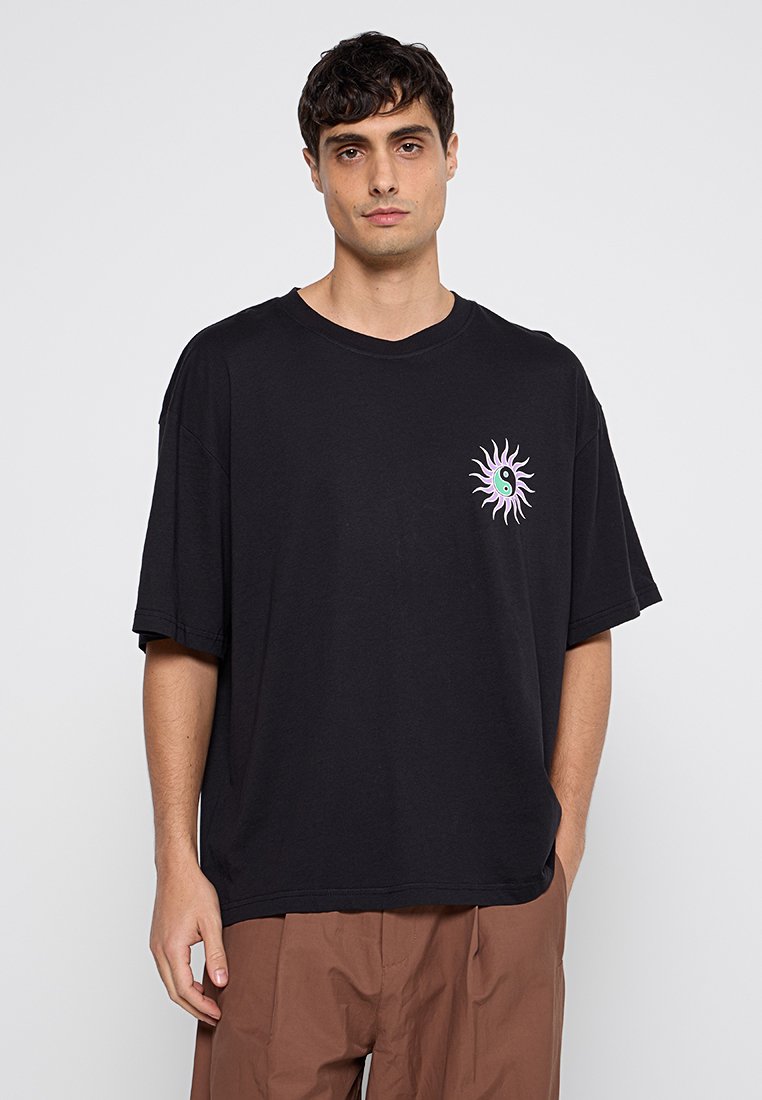T-shirt en coton noir à coupe décontractée, à manches courtes, avec un design coloré de soleil et de yin-yang sur le côté gauche de la poitrine.