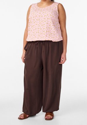 Frau trägt ein ärmelloses pinkes Top mit kleinen gelben Blumen, lockere braune Hosen mit Kordelzug in der Taille und braune Sandalen mit offenen Zehen.