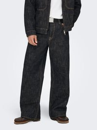 Donker denim jack en broek met wijde pijpen, met contrasterende gouden stiksels, een ritszak en een gestructureerde stofafwerking. Bruine schoenen zichtbaar.