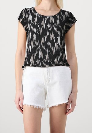 Femme portant un haut à manches courtes noir et gris à motifs et un short en jean blanc à ourlet effiloché, debout sur fond blanc uni.