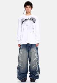 Λευκό πουκάμισο με μακριά μανίκια, με γραφικό και κείμενο "Προστατευτική Ζωτική Δύναμη"· άνετο μπλε τζιν παντελόνι με ανοιχτό γκρι φυλαχτικά μοτίβα· μαύρα παπούτσια.