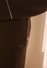 Braunes Set aus langärmligem Oberteil und Hose mit einem kleinen weißen Etikett mit Weinrebenmotiv an der Seitennaht vor beigem Hintergrund.