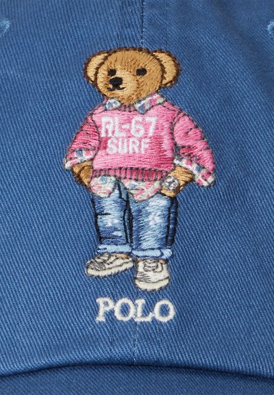Vezen medvedek v rožnatem puloverju z napisom "RL67 SURF," v kavbojkah in teniskah na modri kapici. Spodaj je izšito "POLO."