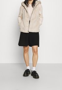 Beige fuskpälsväst med huva, dragkedjeslutning och sidofickor, i kombination med en beige stickad tröja och en svart kjol ovanför knäna, bärs med svarta skor.