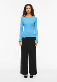 Femme aux cheveux bouclés portant un pull bleu côtelé ajusté avec des poignets et un ourlet à volants, un pantalon large noir et des chaussures noires à bout pointu.