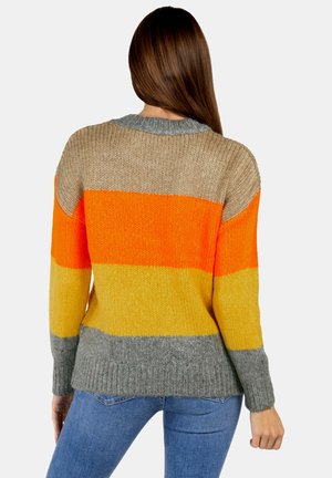 Woman with long brown hair wearing a color-block knit sweater in beige, orange, yellow, and gray, paired with blue jeans, facing away.