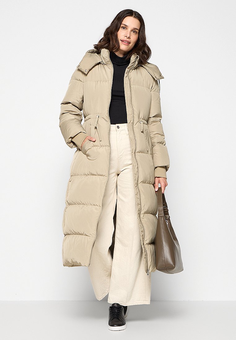 Femme portant un long manteau rembourré beige, un col roulé noir, un pantalon large crème, des chaussures noires, tenant un sac à main marron, se tenant contre un fond blanc.