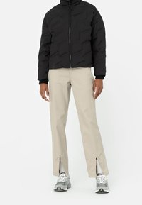 Osoba oblečená v černé zipové bundě, béžových kalhotách s předními rozparky na kotnících, bílých ponožkách a šedých teniskách, stojící na bílém pozadí.