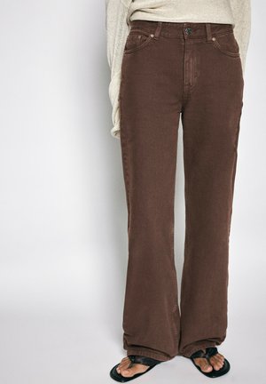 Persoon draagt bruine broek met wijde pijpen, beige trui en zwarte slippers, staand tegen een effen achtergrond.