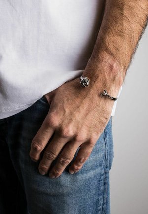 Silver-toned bracelets featuring a floral design and a chain-like pattern, worn on a hand against a light background, above denim jeans.