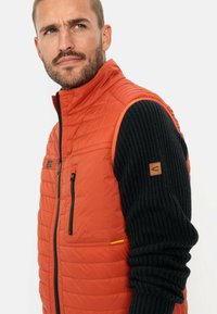 Orangenes isoliertes Weste mit gestepptem Muster, Reißverschlusstaschen und einem schwarzen gerippten Pulloverärmel, der ein Logopatch am Arm hat.