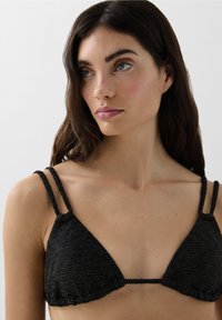 Zwarte driehoek bikini-top van textuurstof met gouden accenten; heeft twee dunne gevlochten bandjes en een gerimpelde rand aan de onderkant.