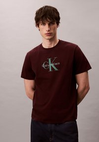Camiseta de algodón burdeos con un gran logo en verde y blanco de "Calvin Klein" con acento de "CK", cuello redondo y mangas cortas.