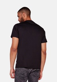 Camiseta de algodón negra con mangas cortas y un cuello redondo, mostrada desde la parte trasera. Presenta una sutil etiqueta de la marca en el dobladillo inferior.