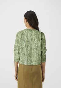 Grønn teksturert bluse med trekvart ermer, med et bølgete mønster. Bak har den en rund hals, kombinert med et beige skjørt.