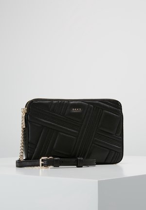 Schwarze, gesteppte DKNY-Umhängetasche mit goldfarbenem Kettenriemen und Reißverschluss, auf weißem Podest vor einfachem Hintergrund präsentiert.